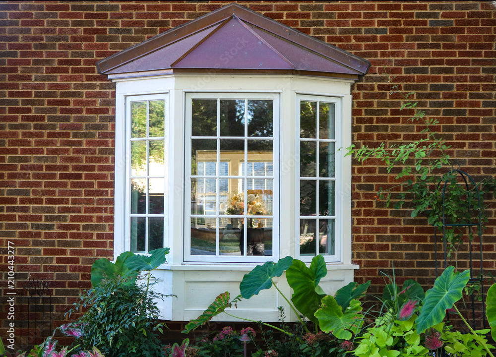 Bay window in a brick house with reflection of trees and view of windows and flowers inside and flowers and elephant ears outside