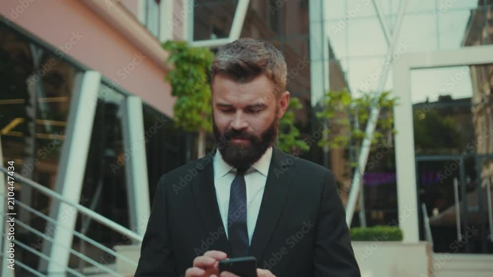 Serious successful business man uses smartphone while walking on the big city business district street office face technology mobile phone suit urban fashion handsome work manager internet outdoors Serious successful business man uses smartphone while walking on the big city business district street office face technology mobile phone suit urban fashion handsome work manager internet outdoors