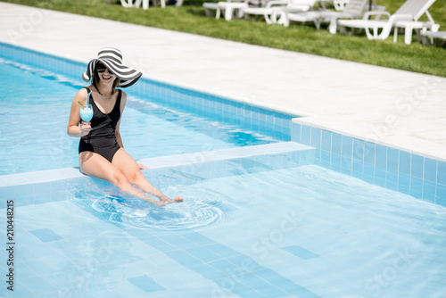 Young woman in black swimsuit and sun hat relaxing with cocktail in the swimming pool outdoors