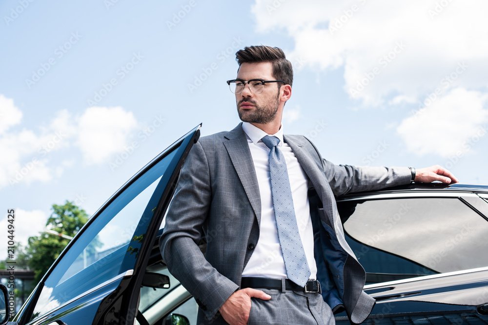 portrait of thoughtful businessman in eyeglasses looking away while standing at car on street