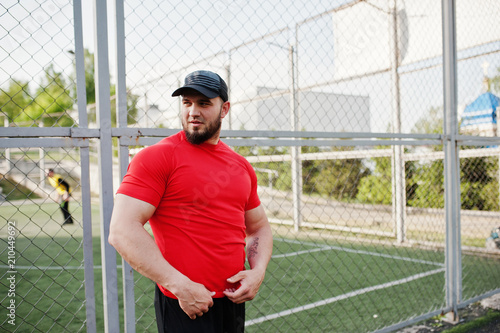 Young brutal bearded muscular man wear on red shirt, shorts and cap at stadium.