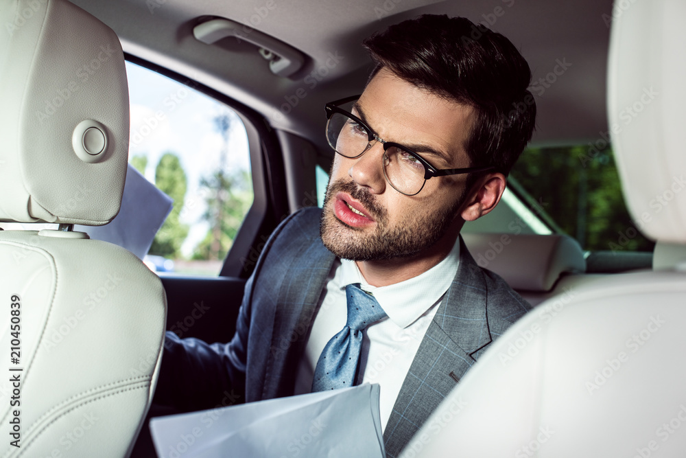 angry young businessman in eyeglasses holding documents while sitting on backseat of car