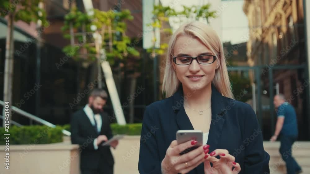 Portrait beautiful young woman with glasses walks down the central city’s street uses phone smile business fashion hand cellphone internet messaging smartphone text happy reading slow motion Portrait beautiful young woman with glasses walks down the central city’s street uses phone smile business fashion hand cellphone internet messaging smartphone text happy reading slow motion