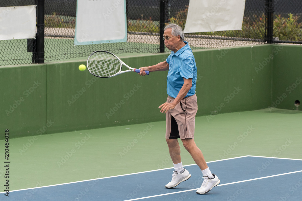 Skilled elderly Chinese tennis player connecting on a forehand ground stroke in a match.