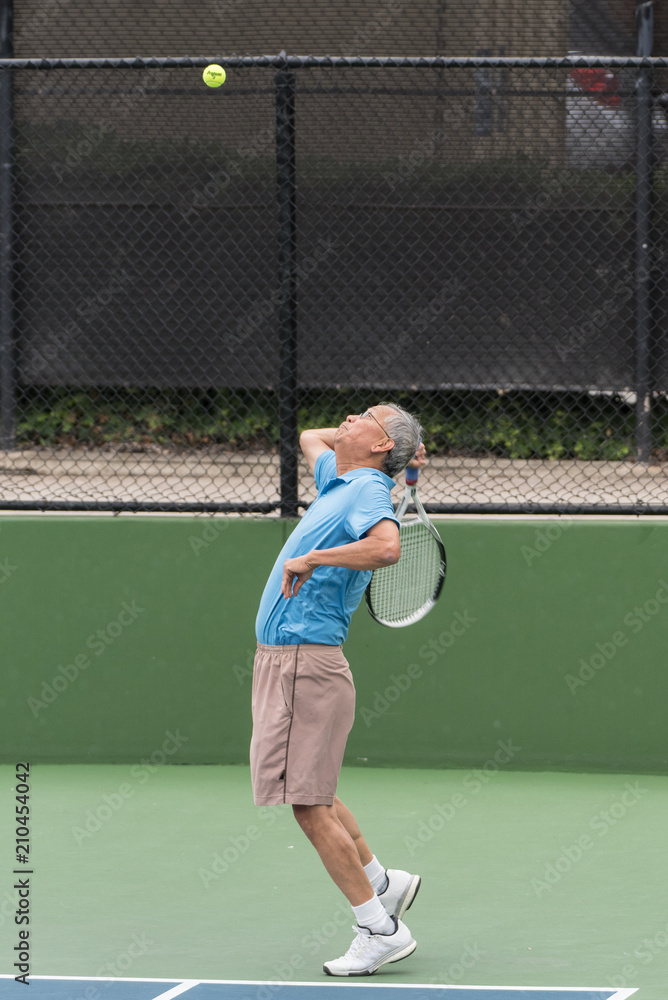 Active Chinese elderly tennis player focused on ball in air with racket back for a power serve