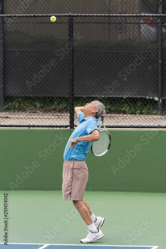 Active Chinese elderly tennis player focused on ball in air with racket back for a power serve © motionshooter