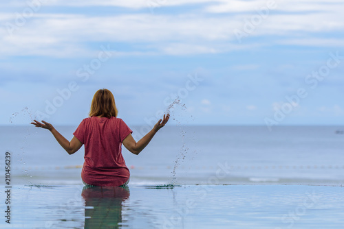 Summer lifestyle traveler woman sitting on the swimmer pool and her hands splashing the pool water  enjoying relaxing on sea beach on summer holiday outdoor vacation trip.