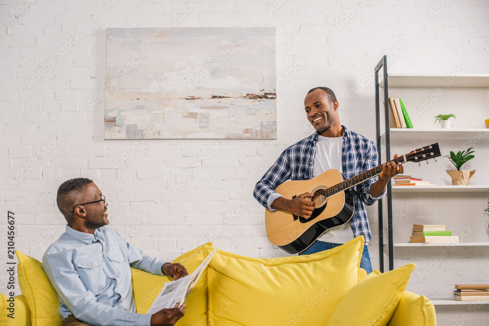 happy senior man with newspaper and smiling adult son with guitar looking at each other at home happy senior man with newspaper and smiling adult son with guitar looking at each other at home