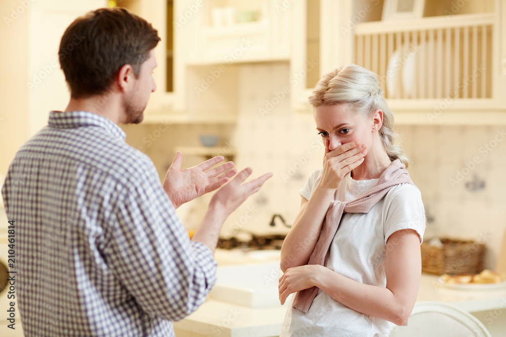 Upset young woman covering her mouth and expressing displeasure while irritated husband talking to her in rude manner Upset young woman covering her mouth and expressing displeasure while irritated husband talking to her in rude manner