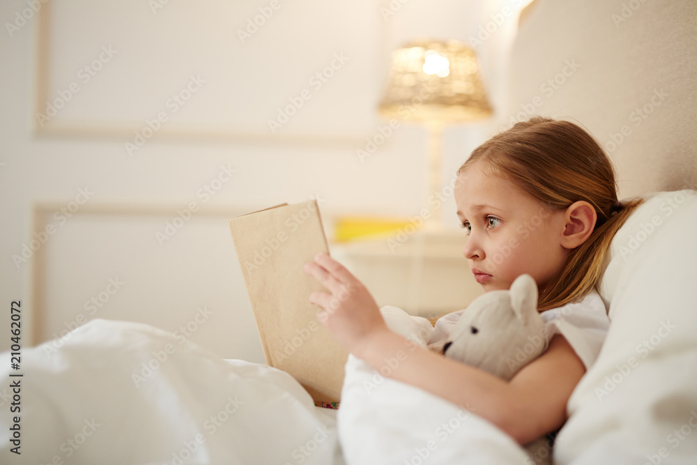 Cute little girl with teddybear lyiing in her bed and reading story tales before sleep Cute little girl with teddybear lyiing in her bed and reading story tales before sleep