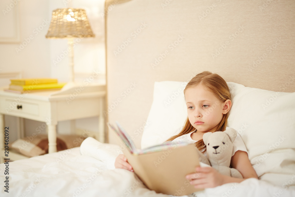 Adorable girl with her favorite toy lying in her bed and reading book of tales in the evening Adorable girl with her favorite toy lying in her bed and reading book of tales in the evening