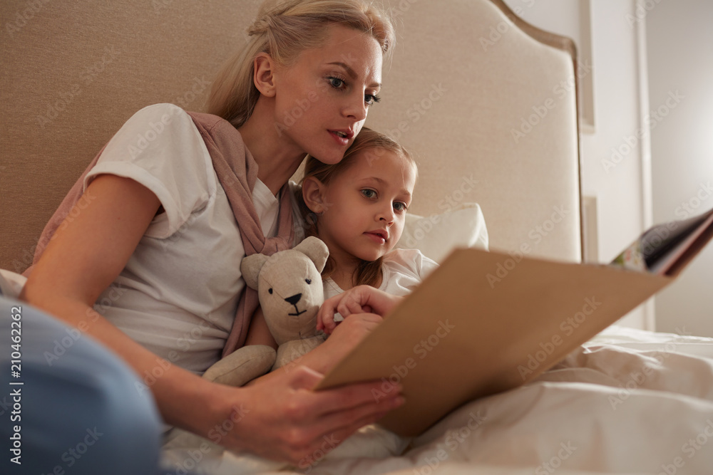 Curious girl and her mother reading book of interesting stories in bed in the evening Curious girl and her mother reading book of interesting stories in bed in the evening