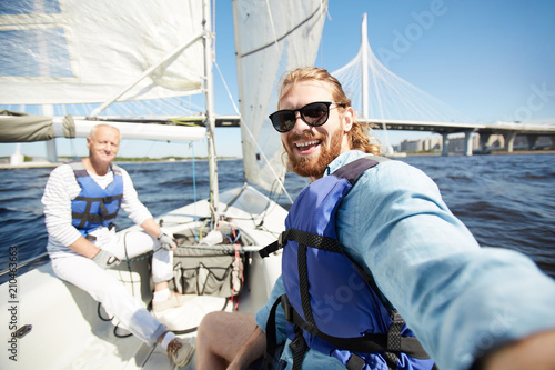Happy young active man in sunglasses and lifejacket making selfie during sailing with senior friend