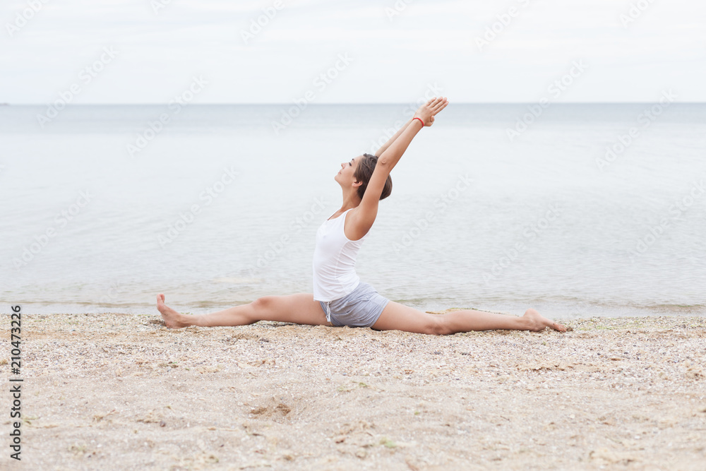 A girl with a short hairstyle dressed in shorts and a white jersey makes a twine on the background of the sea and the beach. Playing sports in nature. Stretching, Yoga pose A girl with a short hairstyle dressed in shorts and a white jersey makes a twine on the background of the sea and the beach. Playing sports in nature. Stretching, Yoga pose