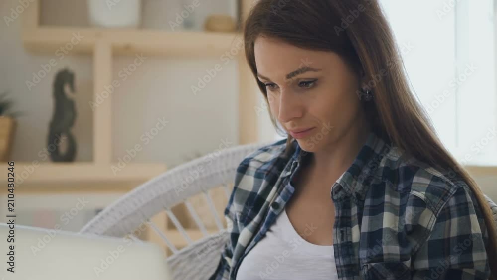 Young beautiful female freelancer with brown hair and in check shirt working on brand-new laptop in white rattan chair with wooden shelves on the background. Indoors. Portrait. Young beautiful female freelancer with brown hair and in check shirt working on brand-new laptop in white rattan chair with wooden shelves on the background. Indoors. Portrait.