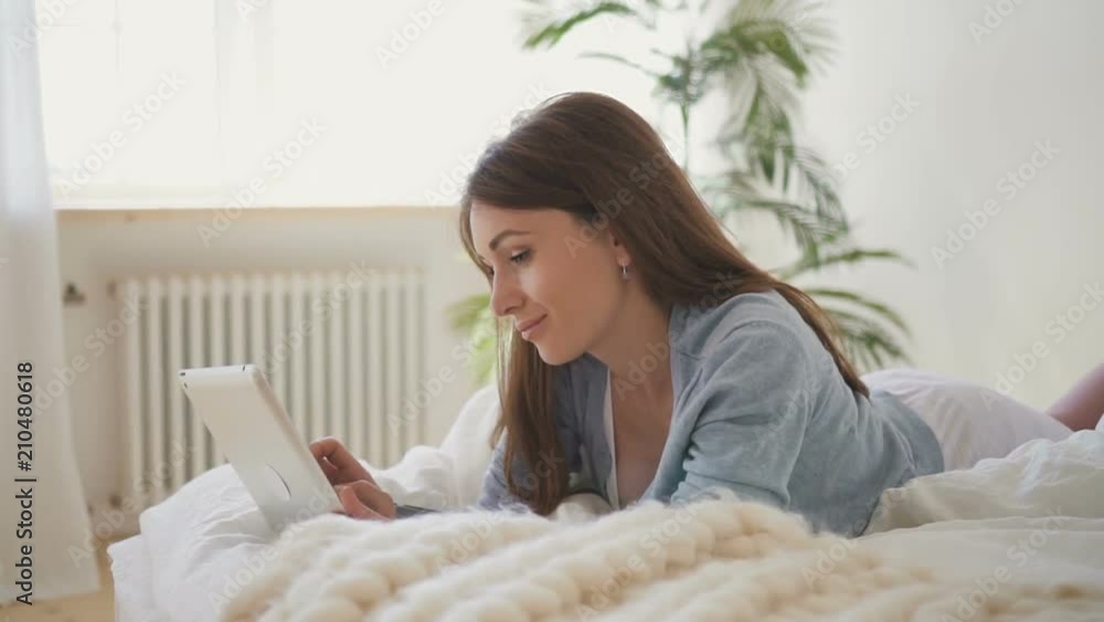Woman in blue t-shirt lying on the bed with white sheets and watching photoes on her brand-new tablet. Indoors. Portrait. Profile. Woman in blue t-shirt lying on the bed with white sheets and watching photoes on her brand-new tablet. Indoors. Portrait. Profile.