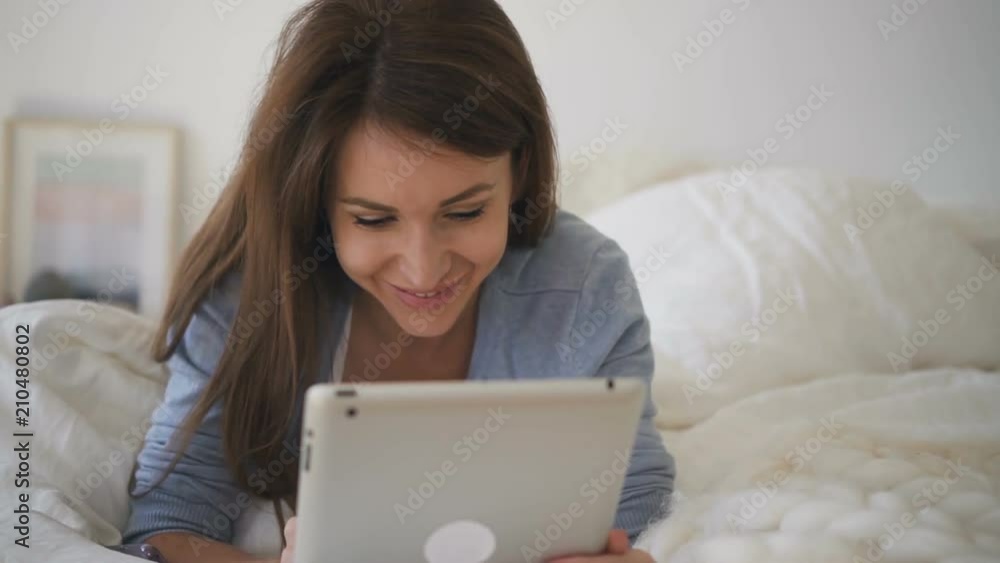 Young woman lying on the bed with white sheets and chatting in social networks on brand-new tablet. Portrait. Indoors. Young woman lying on the bed with white sheets and chatting in social networks on brand-new tablet. Portrait. Indoors.