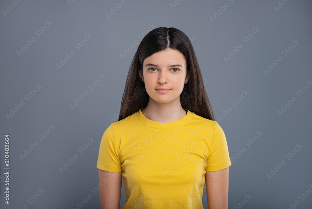 Pretty teen. The portrait of a charming teenage girl in a yellow t-shirt smiling pleasantly while posing against a blue-grey background Pretty teen. The portrait of a charming teenage girl in a yellow t-shirt smiling pleasantly while posing against a blue-grey background
