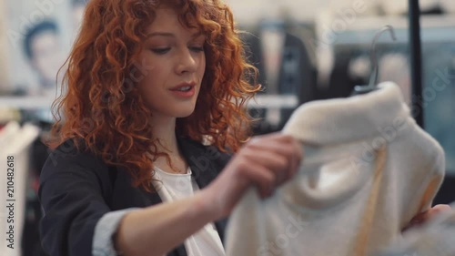 Smiling young woman with curly red hair choose clothes at store hold sweater smile design shopping girl beautiful attractive shop cute happy pretty style curly hair portrait slow motion photo