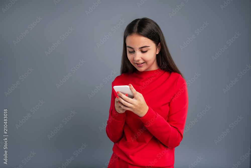 Pleasant chart. Pretty teenage girl sending text messages to her friends while standing isolated on a grey-blue background Pleasant chart. Pretty teenage girl sending text messages to her friends while standing isolated on a grey-blue background