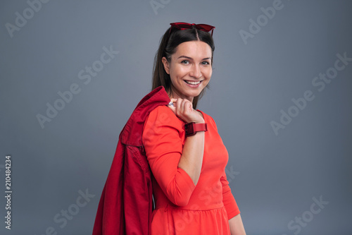 Love fashion. Charming young woman in a red dress posing for the camera and smiling happily while holding a coat
