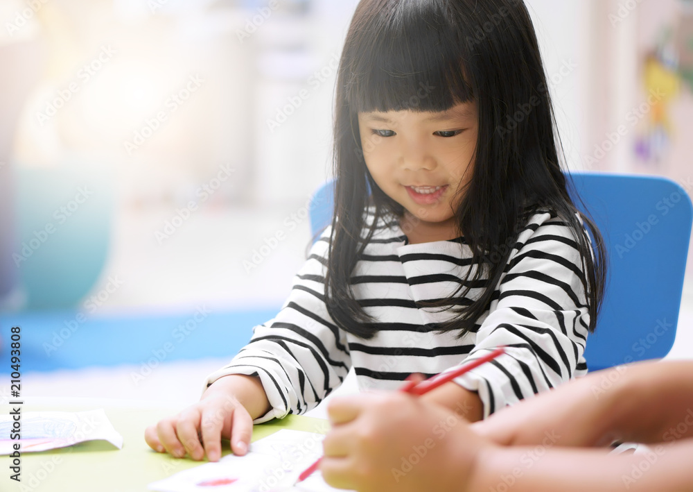 Asian children cute or kid girl smile and learning for coloring or paint on white paper with teacher or mother at nursery or school and kindergarten on soft focus and sunlight on window Asian children cute or kid girl smile and learning for coloring or paint on white paper with teacher or mother at nursery or school and kindergarten on soft focus and sunlight on window
