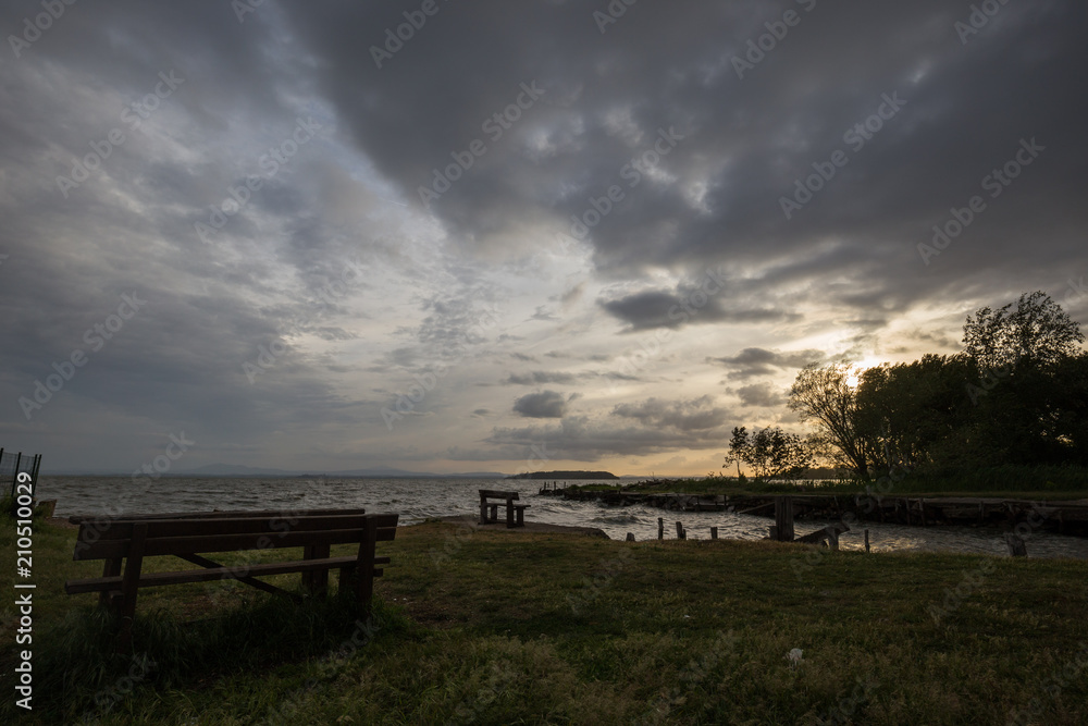 Wooden sitting benches on a lake shore at sunset, beneath a moody, cloudy sky