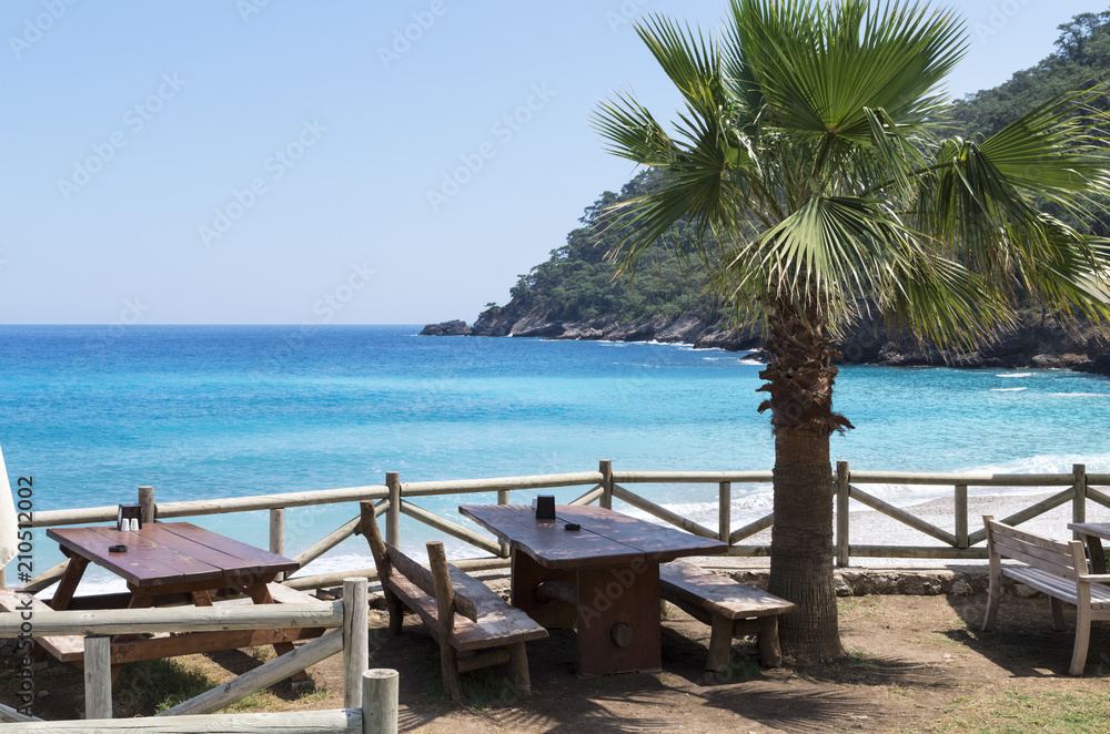 The concept of summer holidays or vacation on the seacoast; Summer open cafe on the sandy beach of the Mediterranean Sea; Blue sea, sandy beach, green palm trees and cafe with open terrace