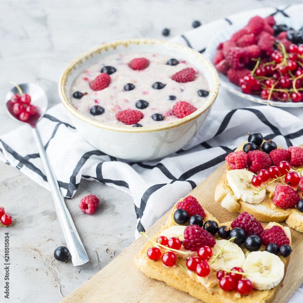 A table with a healthy summer breakfast - oatmeal with berries, currants and raspberries, and a sandwich with peanut butter, banana and various berries A table with a healthy summer breakfast - oatmeal with berries, currants and raspberries, and a sandwich with peanut butter, banana and various berries