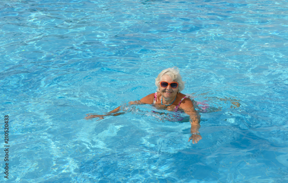 Aged woman is swimming in bright blue water of pool.