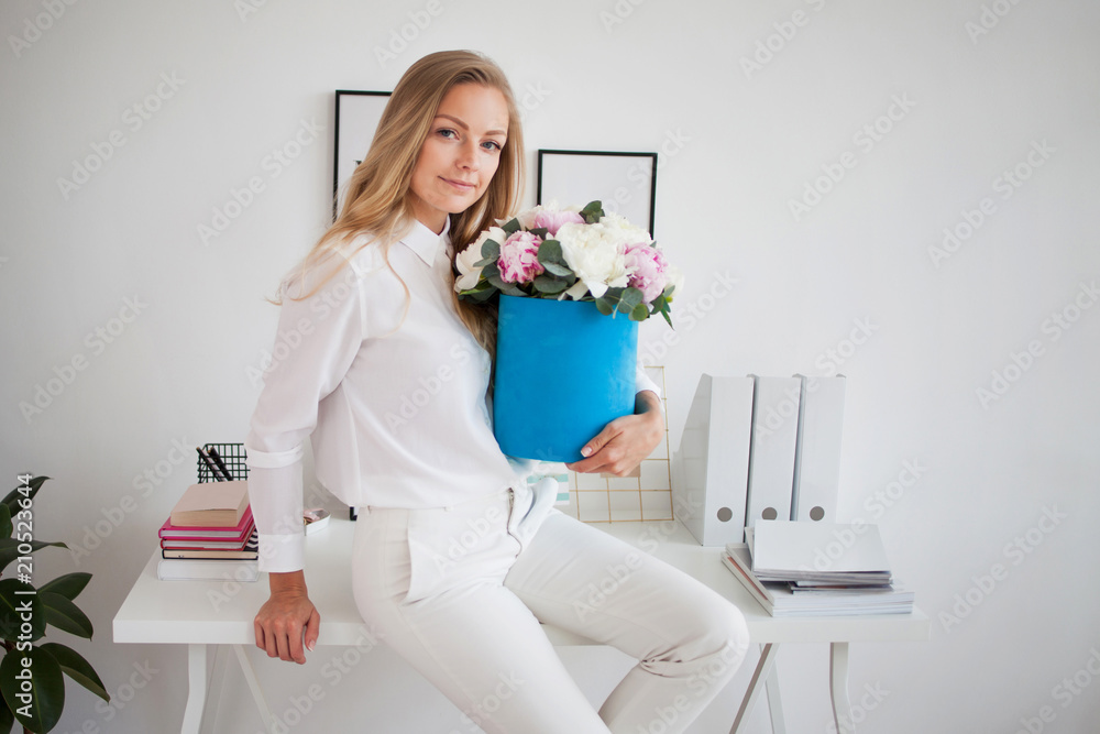 Stylish young blonde in office, white shirt and pants. Holds a luxurious bouquet of peonies in a blue hat box. Stylish young blonde in office, white shirt and pants. Holds a luxurious bouquet of peonies in a blue hat box.