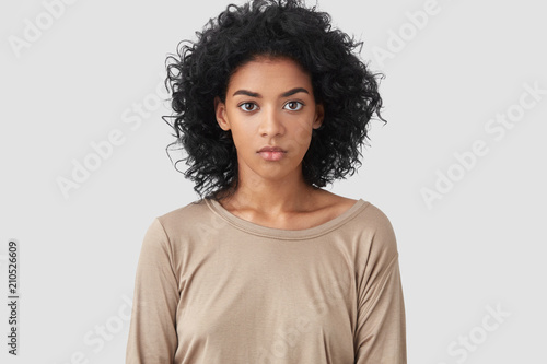Indoor shot of serious dark skinned female freelancer has Afro hairstyle, pleasant appearance, dressed in beige casual sweater, works distantly at home, enjoys domestic atmosphere. Ethnicity concept