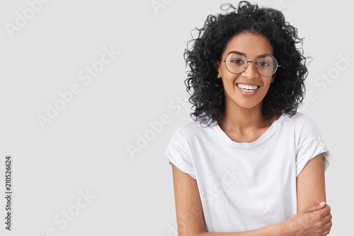 Waist up portrait of beautiful young dark skinned female with positive expression, has Afro hairdo, feels happy after successful bargain, wears big spectacles, isolated over white background