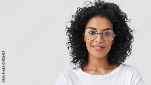 Close up portrait of curly female adult has charming smile, curly dark hair, wears big glasses, satisfied as finished domestic work earlier, being successful designer or architect, has talent photo