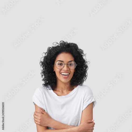 Glad female student has Afro hairstyle, broad smile, keeps hands crossed, wears glasses, happy to spend evening with family, wears t shirt in one tone with background. People and emotions concept