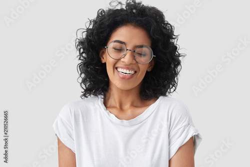 Headshot of pretty African American female being in high spirit, keeps eyes closed, smiles at funny small child, expresses sincere emotions, wears casual white t shirt, isolated over blank wall
