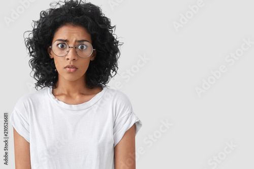 Frightened stupefied frustrated African American female looks in bewilderment with wide opened eyes, afraids of something, wears big spectacles, isolated over white background with copy space.