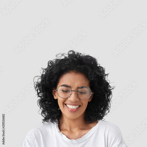 Canvastavla Vertical shot of overjoyed mixed race female laughs as watches comedy serial, has toothy smile, shows white perfect teeth, closes eyes from laughter, has crisp hair and dark skin