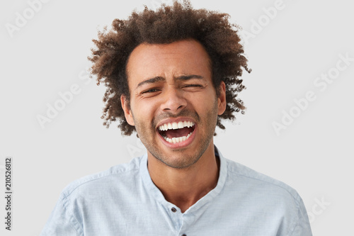 Optimistic glad African American male has broad smile, laughs at funny story, expresses positive emotions, being in good mood, isolated on white concrete studio wall. Happy mixed race young man