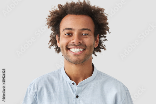 Headshot of handsome mixed race young man with shining smile, looks positively at camera, has white perfect teeth, stands against white background. People, lifestyle, emotions and fun concept photo