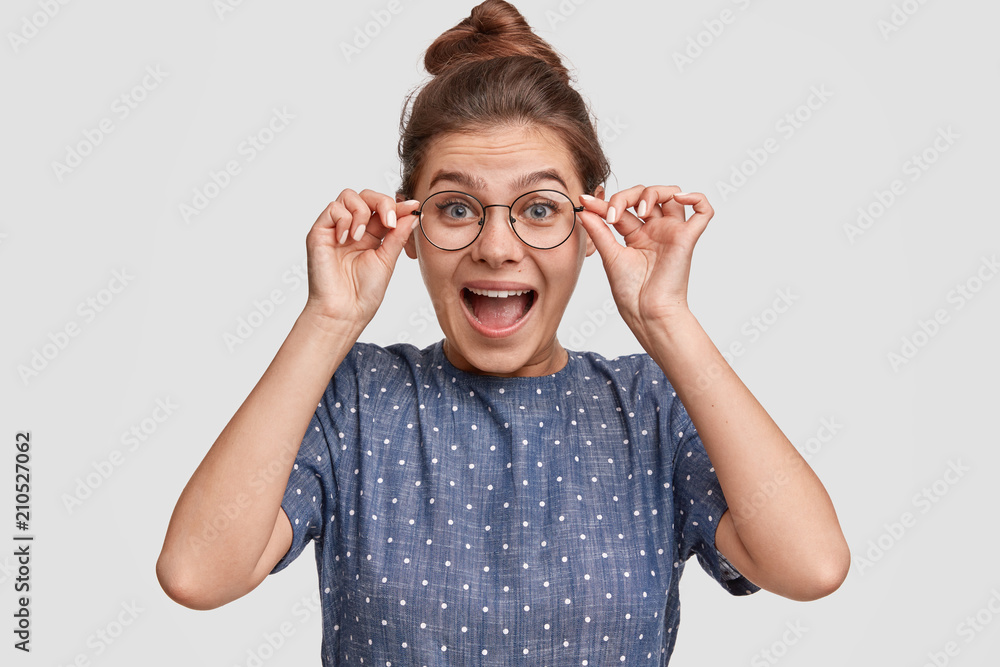 Studio shot of amazed beaitiful female recieves desirable thing on birthday, stares through spectacles, keeps hands on rim, being in high spirit, poses over white background. Emotions concept Studio shot of amazed beaitiful female recieves desirable thing on birthday, stares through spectacles, keeps hands on rim, being in high spirit, poses over white background. Emotions concept