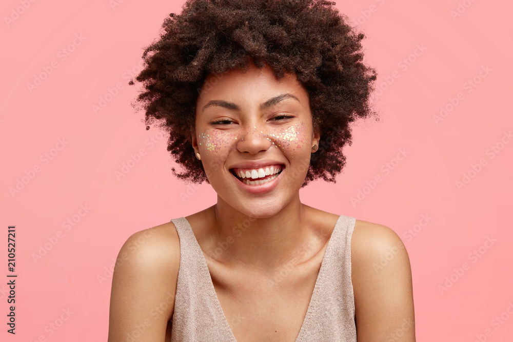 Attractive woman with pleased expression, smiles joyfully, being glad satisfied, isolated on pink background, has glitter on face, dressed in casual t shirt. People, ethnicity and beauty concept Attractive woman with pleased expression, smiles joyfully, being glad satisfied, isolated on pink background, has glitter on face, dressed in casual t shirt. People, ethnicity and beauty concept
