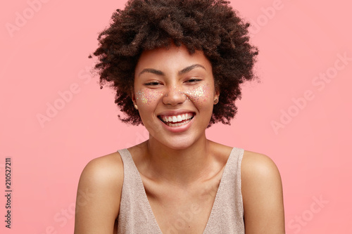 Attractive woman with pleased expression, smiles joyfully, being glad satisfied, isolated on pink background, has glitter on face, dressed in casual t shirt. People, ethnicity and beauty concept