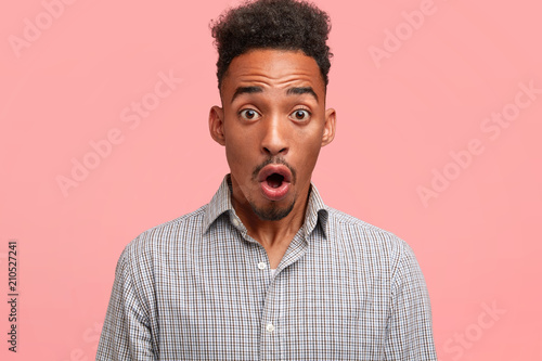 Horizontal shot of surprised dark skinned male student being stupefied to recieve bad mark for project work, dressed in checkered shirt, isolated on pink wall. African American man expresses shock