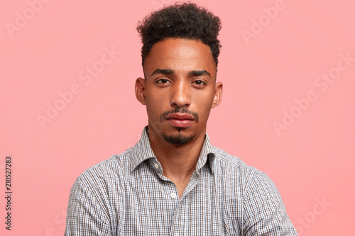 Horizontal shot of serious mixed race attractive self assured businessman with specific appearance, looks directly at camera, ready for negotioation with partners, dressed in elegant checkered shirt