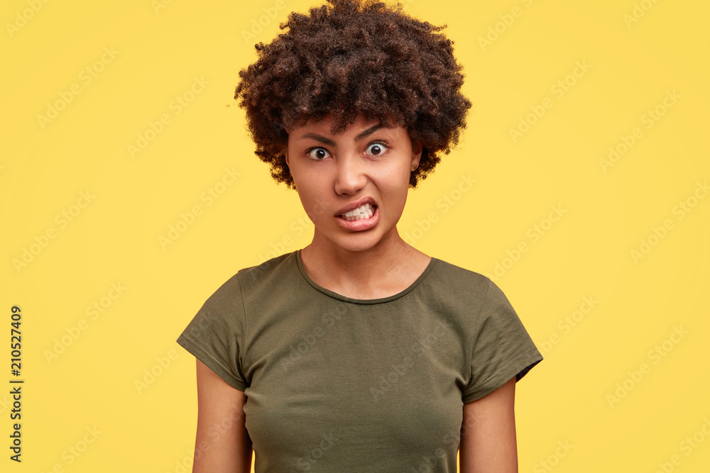 Portrait of annoyed Afrrican American female clenches teeth with irritation, has quarrel with husband, raises eyebrows in anger, isolated over yellow background. People and negative emotions concept Portrait of annoyed Afrrican American female clenches teeth with irritation, has quarrel with husband, raises eyebrows in anger, isolated over yellow background. People and negative emotions concept