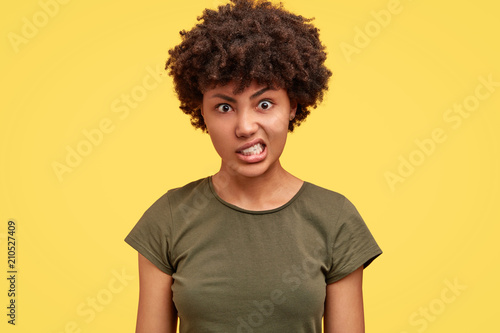 Portrait of annoyed Afrrican American female clenches teeth with irritation, has quarrel with husband, raises eyebrows in anger, isolated over yellow background. People and negative emotions concept