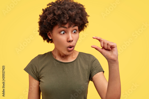Surprised beautiful African American female stares with bugged eyes and makes tiny gesture, shows how much attention she recieves from boyfriend, poses against yellow background. It`s too small photo