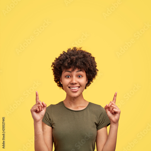 Joyful pretty African American female indicates with both fore fingers up, has friendly smile, dark skin, curly hair, shows blank space against yellow background. People, advertisement concept