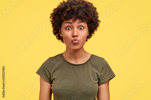 Isolated shot of pretty dark skinned woman makes grimace, keeps lips round, expresses surprisement, has appealing appearance and Afro hairstyle, isolated on yellow wall. Facial expressions concept